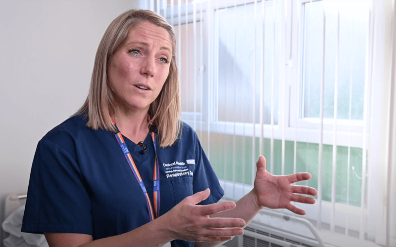 A screen capture of webinar where a healthcare professional in a blue uniform labeled "Oxfordshire Respiratory Team" gestures with her hands, speaking in a well-lit room with white blinds covering the windows.