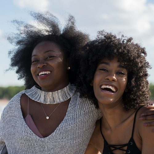 Two people are smiling and embracing outdoors. They appear joyful, with a clear blue sky and trees in the background.