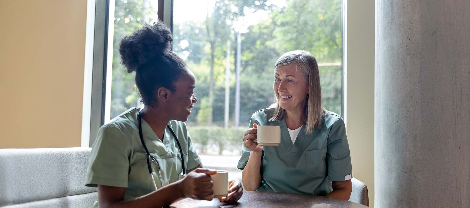A young black nurse and a middle aged blonde nurse sat talking and having a cup of tea on their lunch break.