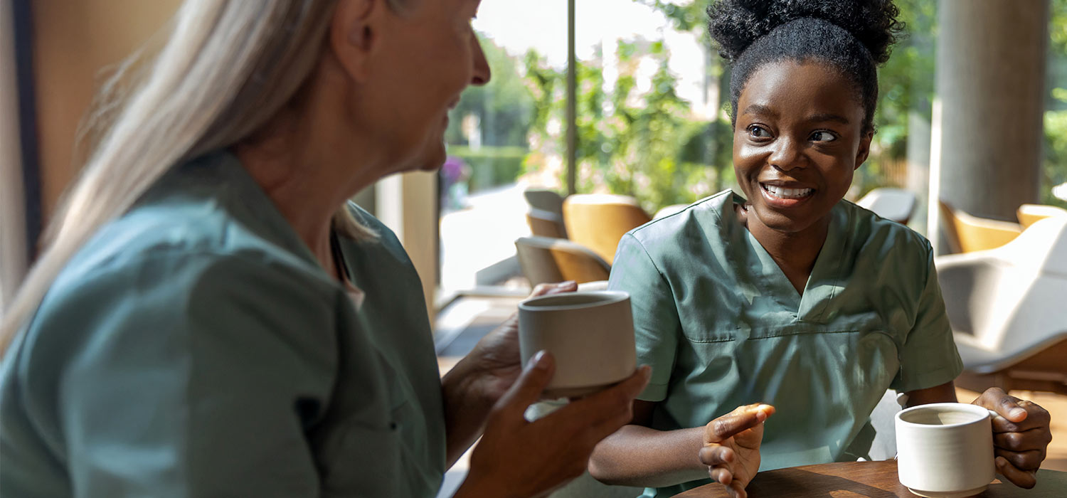 A young black nurse and a middle aged blonde nurse sat talking and having a cup of tea on their lunch break.