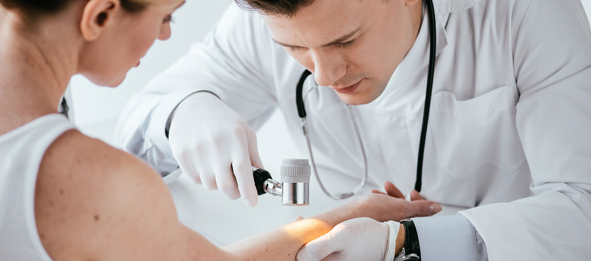Dermatoscope examining skin on a person's arm; a gloved hand holds the device in a medical setting.