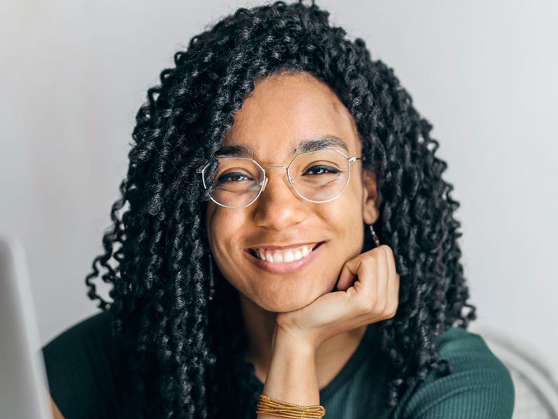 A woman with black tight ringlet curls, wearing silver framed glasses sits with her chin on her palm as she smiles at the camera.