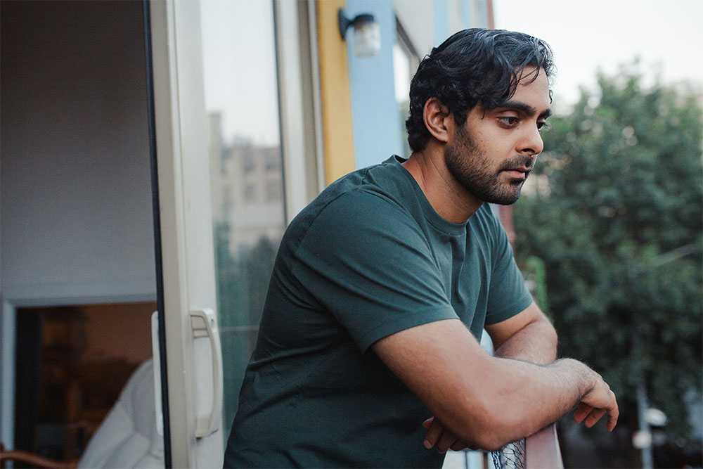 Man in a green shirt leans on a balcony railing, gazing thoughtfully into the distance. Trees and a building are visible in the background, with part of an open door on the left.