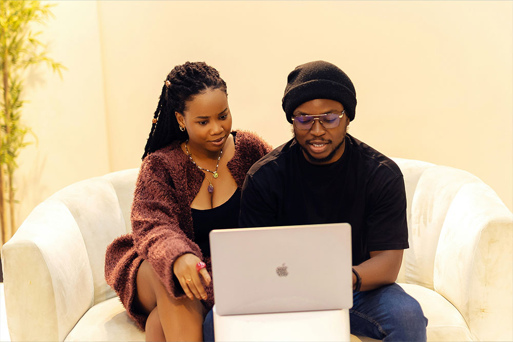 Two people sit together on a white sofa, focusing on a laptop screen. A potted plant is visible in the background, enhancing the warm, inviting atmosphere.