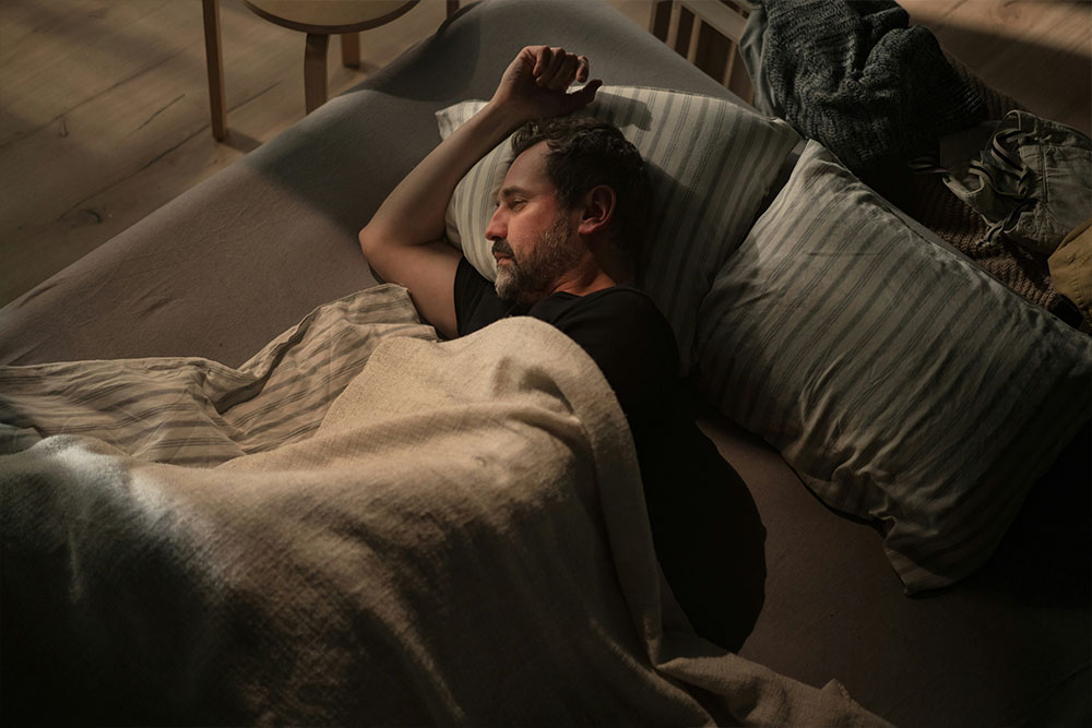 A person sleeps on a bed, covered with a blanket and resting on striped pillows, in a dimly lit room with wooden flooring and furniture.