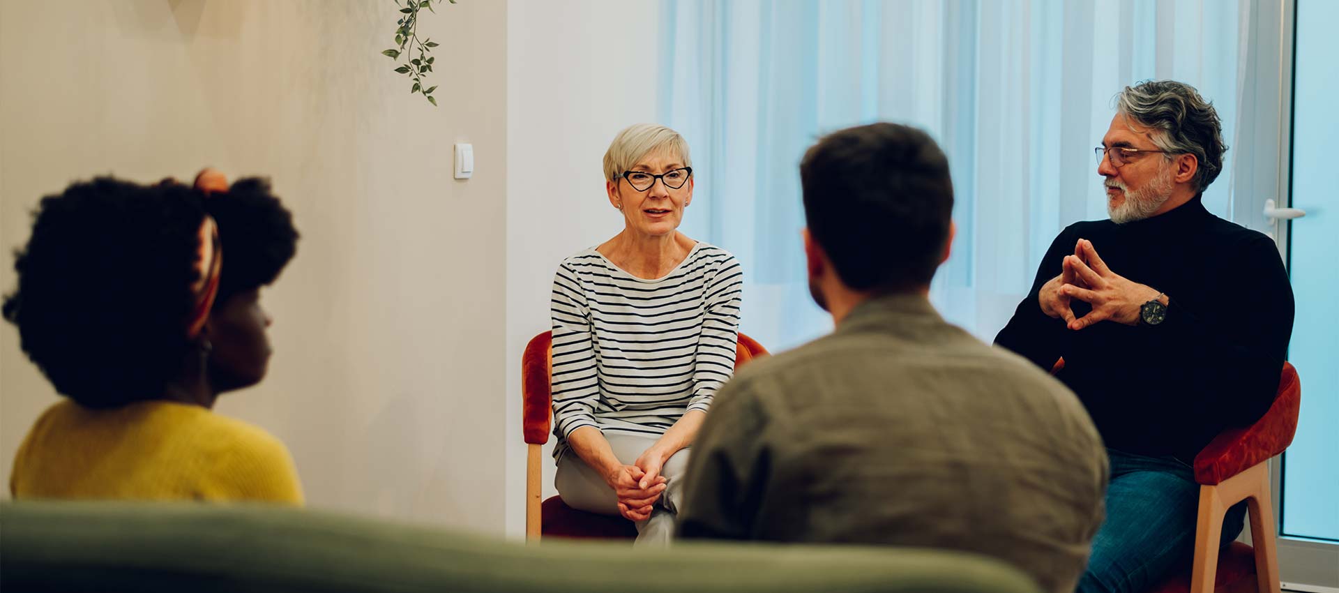 A group of four people engage in a discussion, seated in a circle within a softly lit room with a plant and light curtains in the background.