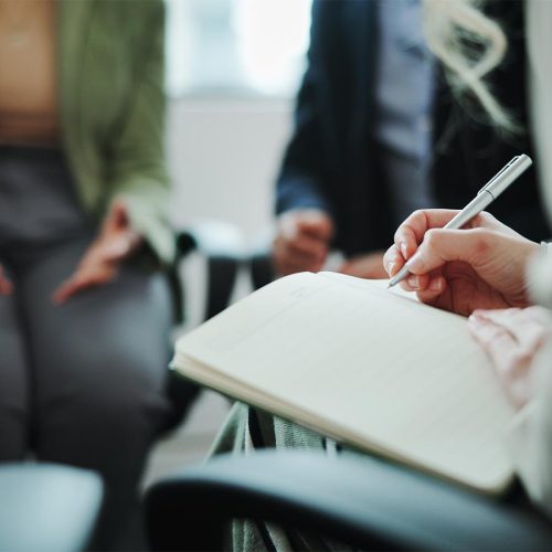 A person holding a notepad and pen writing down notes with a blurred background of a meeting.