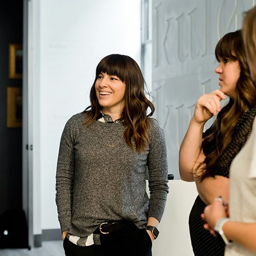 A person with long hair stands smiling, engaged in conversation with another individual. They are in a modern indoor setting with minimalistic decor and soft lighting.