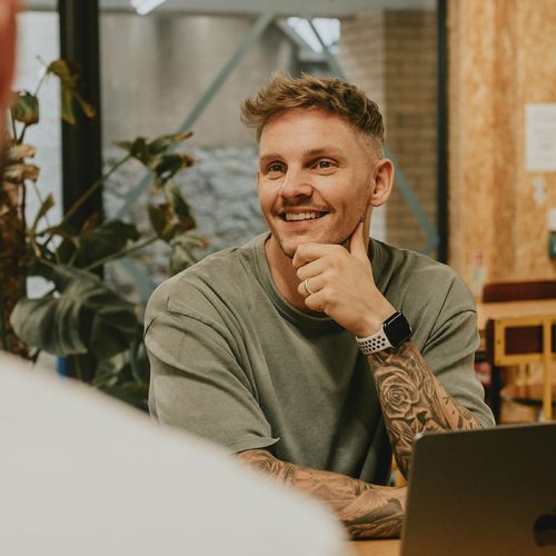 A young man with tattoos down both his arms sits at a desk with a silver laptop infront of him while he talks to another man sat opposite him.