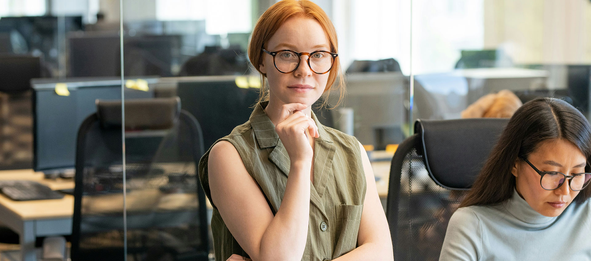 Woman standing thoughtfully with hand on chin, wearing glasses and a sleeveless top in an office environment with colleagues working at desks in the background.