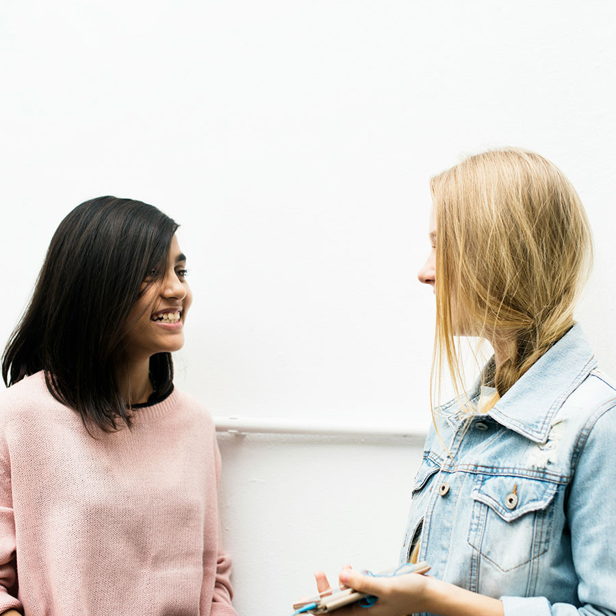 Two people engage in a friendly conversation indoors. One holds a clipboard and pen, while the other smiles. They stand against a plain white wall.