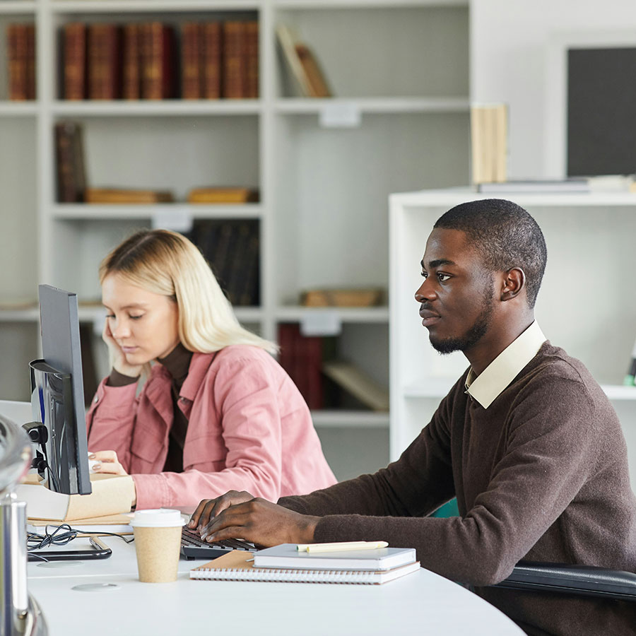 Two people sit at a desk in an office, focused on their computer work. Shelves with books are visible in the background, and a coffee cup rests on the desk.