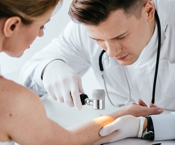 A doctor examines a patient's skin with a dermatoscope, focusing intently. The patient is seated across a desk in a clinical setting, wearing a white coat and rubber gloves.