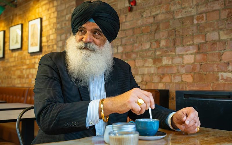 Man wearing a turban and suit stirring a blue cup at a wooden table in a cafe with exposed brick walls and framed pictures.