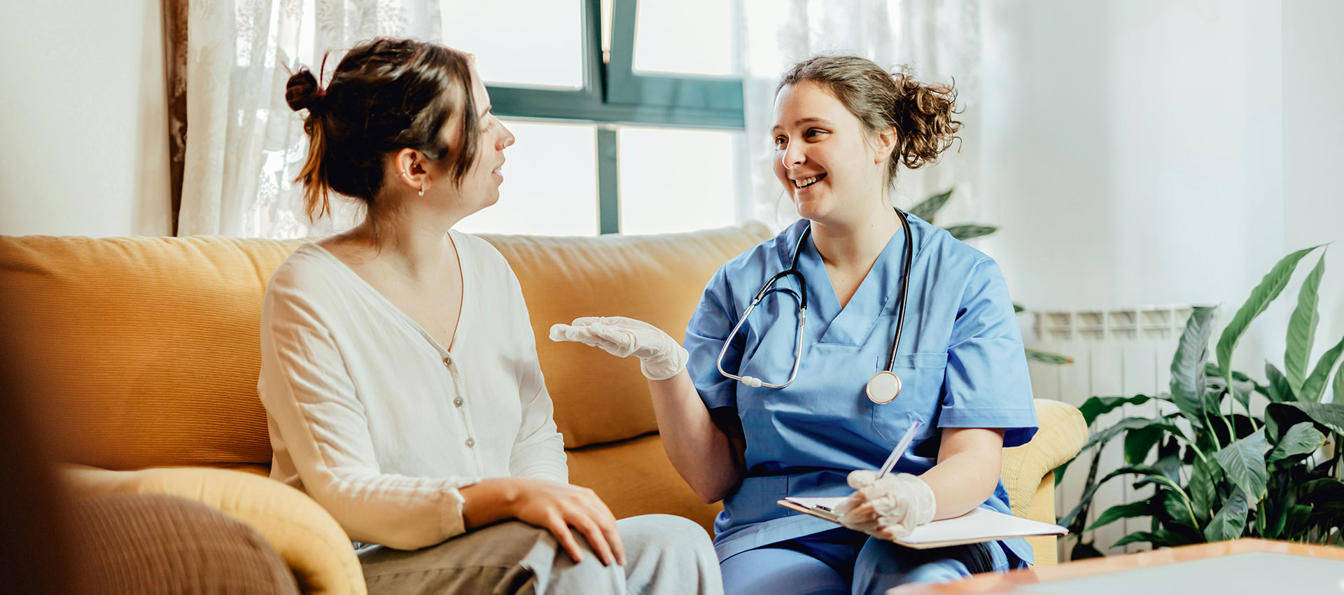 A healthcare professional, in blue scrubs, sits on a couch smiling and conversing with a woman in a living room setting, with plants and soft lighting in the background.