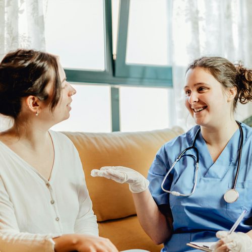 A healthcare professional in blue scrubs and gloves converses with a seated woman in a white top. They are indoors, with light streaming through a window behind them.