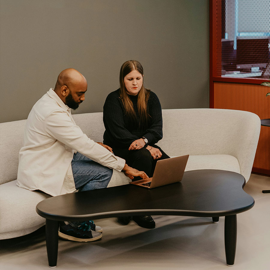 Two people sit on a cream sofa, engaging with a laptop on a black, abstract-shaped coffee table in a modern office setting.