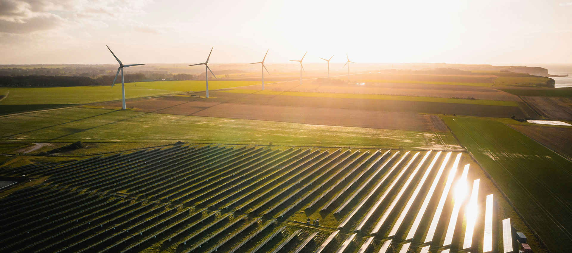 Wind turbines stand in a distant field as the sun sets, casting a warm glow over rows of solar panels and expansive green farmland.