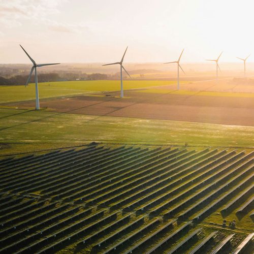 Wind turbines stand in a line on a vast, sunny landscape with fields and solar panels in the foreground, under a clear sky.