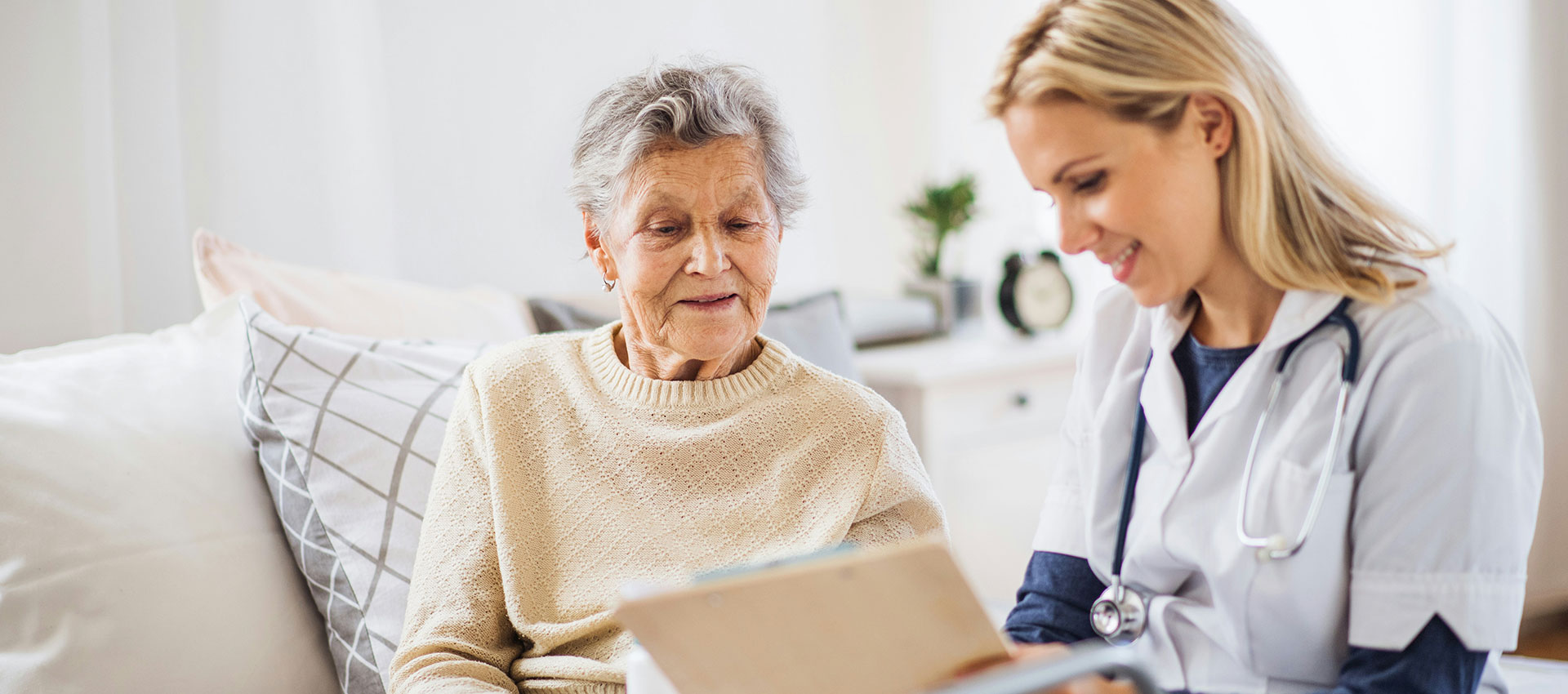 A healthcare professional consults an elderly woman, reviewing documents together in a cozy, well-lit room with cushions and a clock visible in the background.