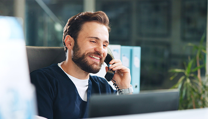 A man is smiling while talking on a phone, sitting at a desk in a modern office environment, with sunlight streaming through a window.
