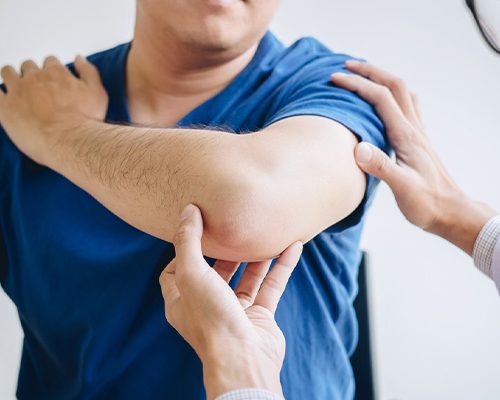 A person in a blue shirt, with an arm bent at the elbow, is examined by another person’s hands, suggesting a clinical or therapeutic setting.