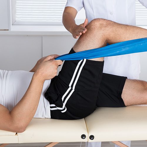 A person receives physiotherapy, using a blue resistance band to stretch their leg while a therapist assists. They lie on a treatment table in a clinical setting.