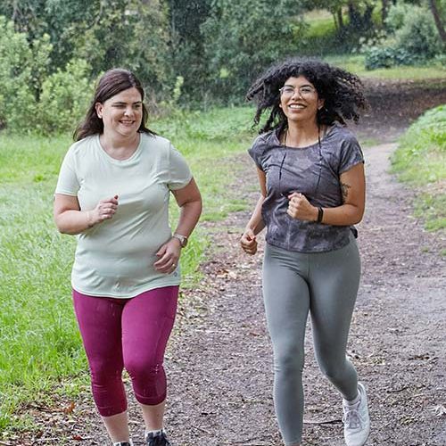 Two women jog along a gravel path in a lush, green park, smiling and enjoying their exercise. Trees and bushes line the path, creating a serene outdoor environment.
