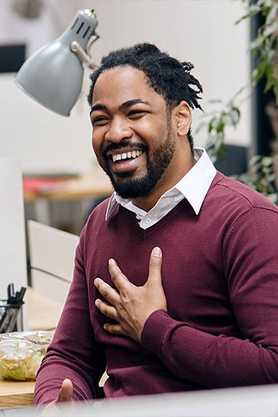 A person in a maroon sweater gestures with a hand on their chest, smiling warmly. They are in an office setting with a desk lamp and plants visible in the background.