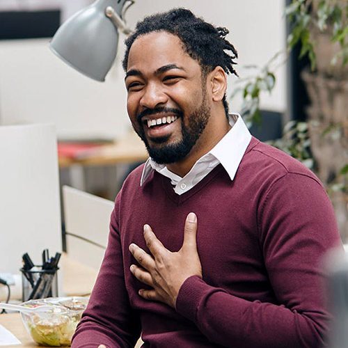 A person in a maroon sweater gestures with a hand on their chest, smiling warmly. They are in an office setting with a desk lamp and plants visible in the background.