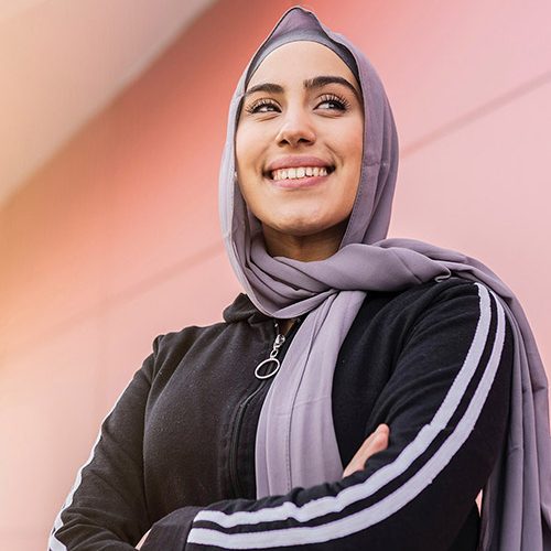 A person in a black jacket and lavender hijab smiles confidently with arms crossed. The individual stands against a pink wall with diagonal lines in the background.