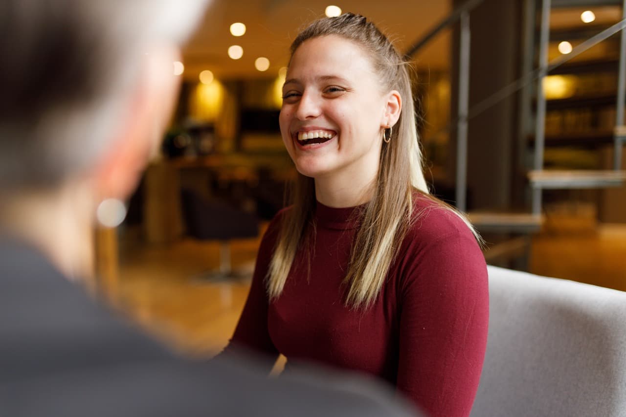A person in a maroon top sits and smiles brightly, engaging in conversation with another person, in a warmly lit, modern indoor setting with blurred background elements.