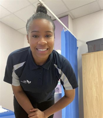 A person in a navy uniform smiles while leaning forward slightly. They are in a medical examination room with a blue privacy curtain and a wooden cupboard in the background.