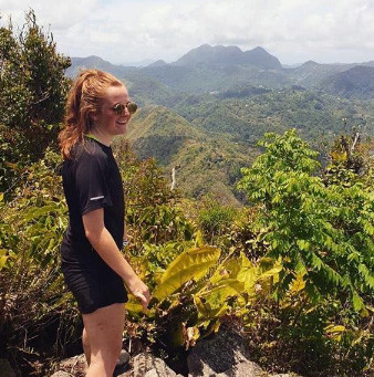 A person wearing sunglasses stands on a mountain ledge, surrounded by lush greenery, overlooking a vast landscape of rolling hills under a partly cloudy sky.