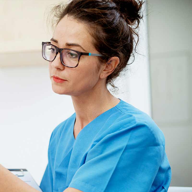 Close up of a woman wearing blue scrubs and black glasses looking at something off camera.