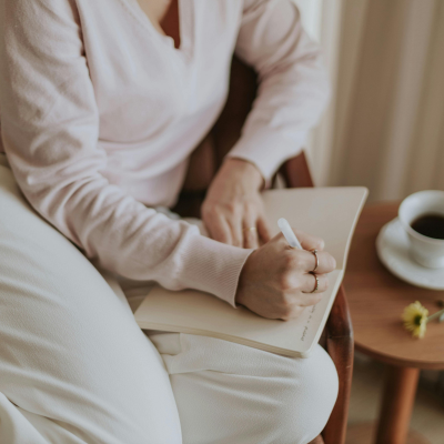 A person sitting comfortably and writing in a notebook, with a cup of coffee on a small table beside them.