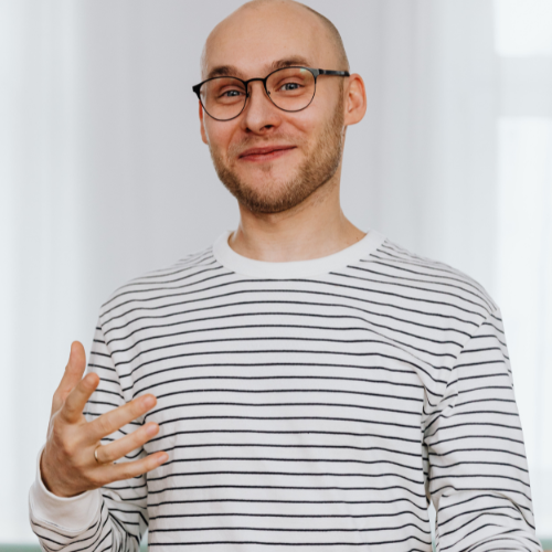 A person wearing a striped long sleeve top gesturing with their hands while speaking, standing in a bright indoor setting.