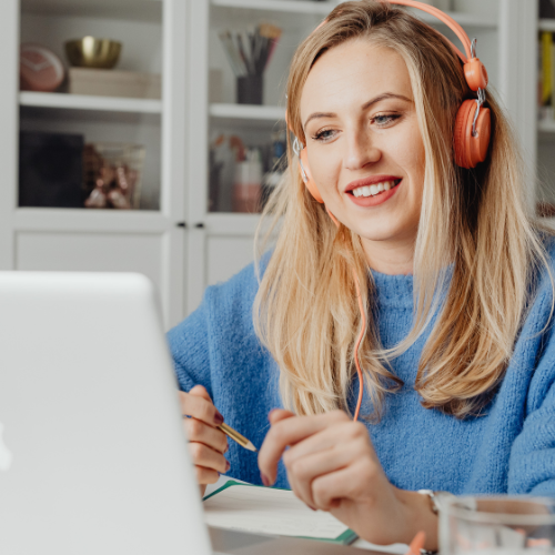 A person wearing orange headphones sitting at a laptop, writing in a notebook in a bright home workspace.