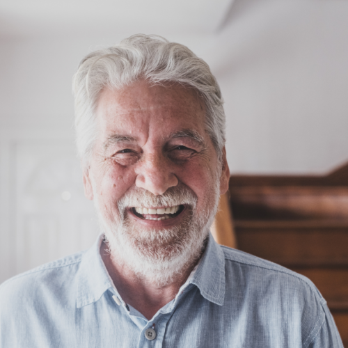 An elder man with grey hair stands indoors in a softly lit home environment, with a wooden staircase and hallway visible in the background.